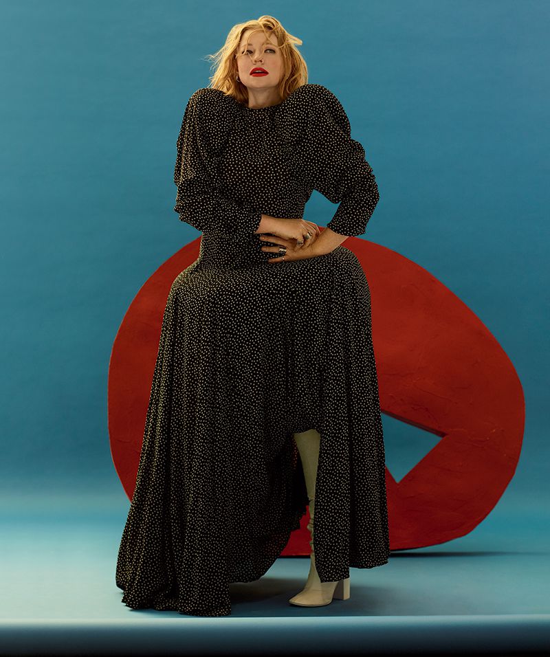 A fair-skinned woman with blonde hair poses in a polka dot dress in front of a red abstract sculpture on a blue backdrop.