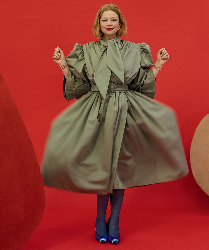 Adult female in studio poses in an olive-green dress and blue shoes against a red background with geometric shapes.