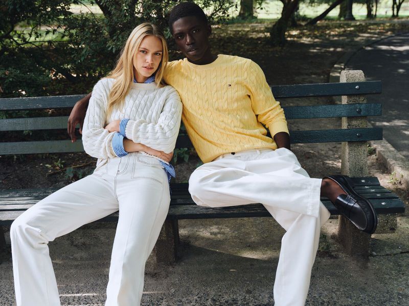 A stylish couple poses on a park bench. The woman wears a white sweater and jeans, while the man wears a yellow sweater and white pants.