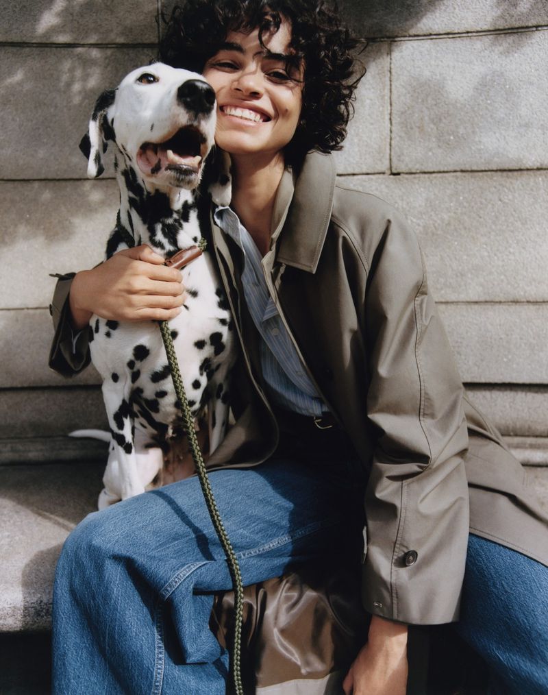 Woman smiling with Dalmatian dog on leash, wearing trench coat and jeans, sitting against a stone wall in natural daylight.