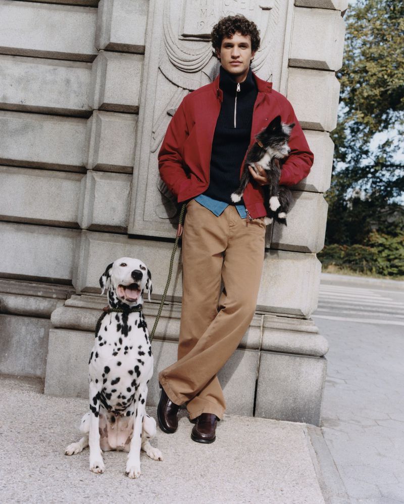 A man standing in a park with a Dalmatian dog and a cat, wearing a red jacket and tan pants, with trees and architectural details in background.