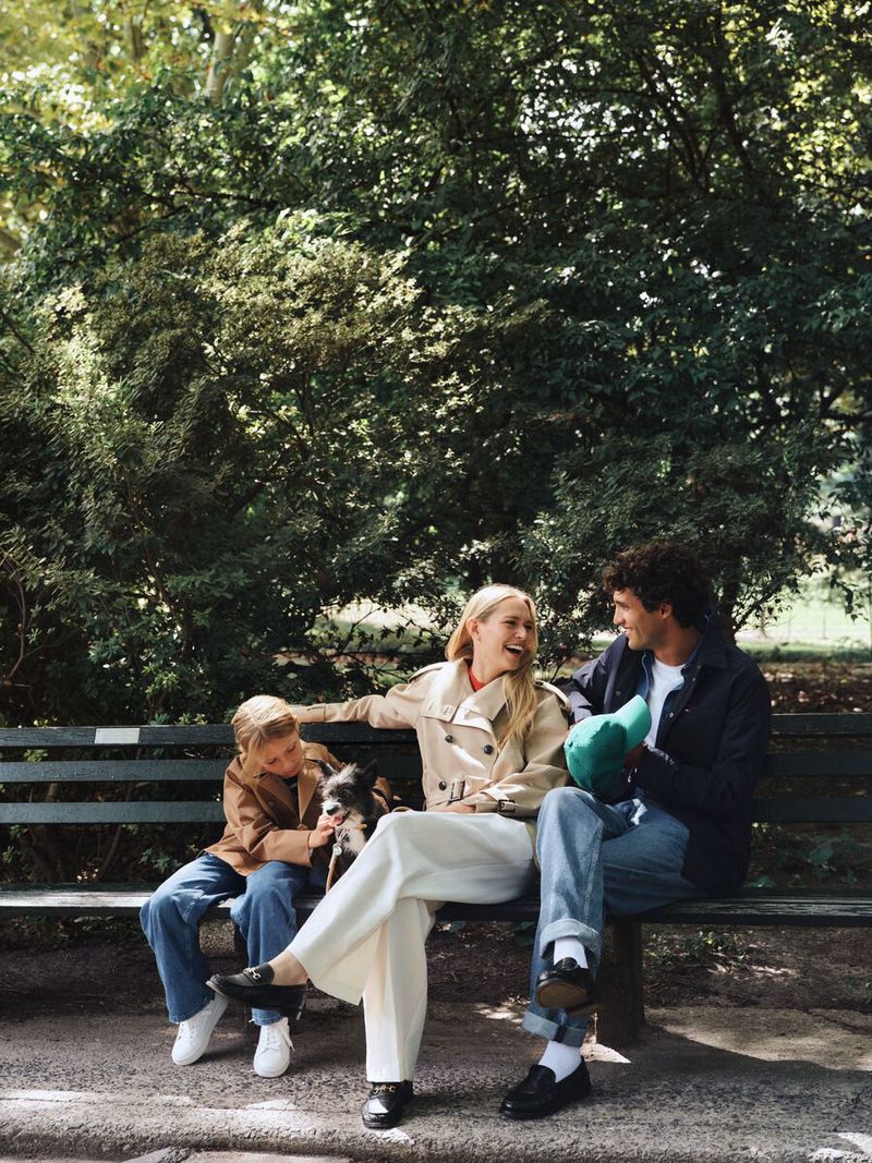 Family sits on a park bench with their dog. A blonde woman smiles at a man holding a hat. A child pets the dog with trees behind them.