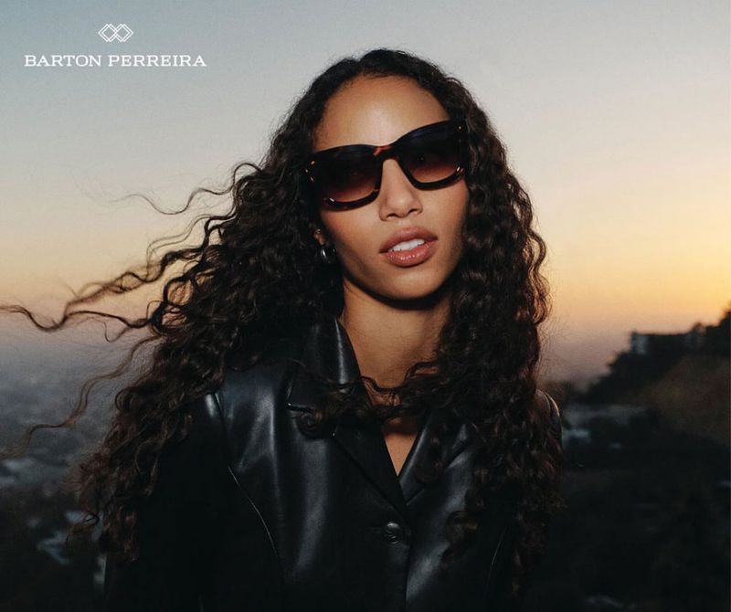 Woman wearing dark sunglasses and black jacket with long curly hair against a sunset sky background for Barton Perreira