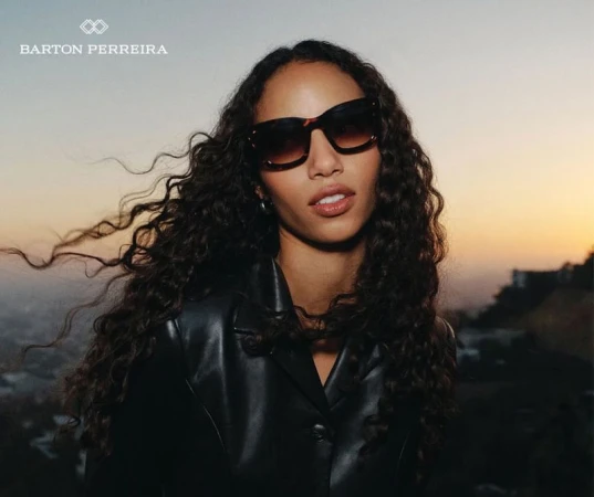 Woman wearing dark sunglasses and black jacket with long curly hair against a sunset sky background for Barton Perreira