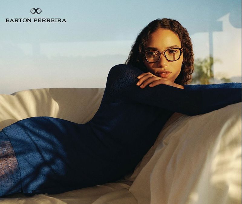 Woman in a navy dress reclines on a white surface, wearing tortoise-shell glasses, in a bright, naturally lit setting with neutral background.