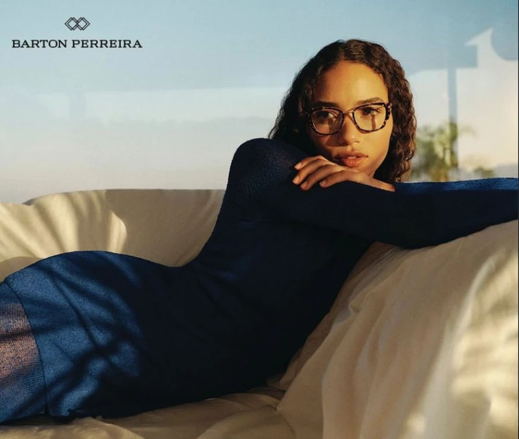 Woman in a navy dress reclines on a white surface, wearing tortoise-shell glasses, in a bright, naturally lit setting with neutral background.