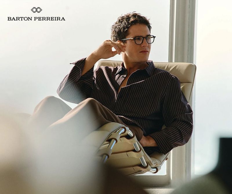 Adult male model with dark curly hair sits in a chair wearing glasses in a bright space; the Barton Perreira logo is visible.