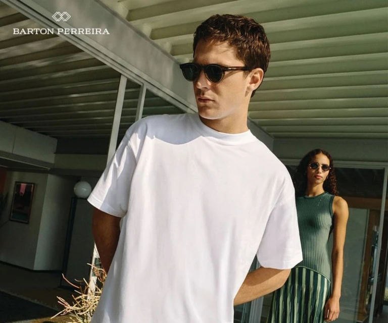 A fashionable couple outside a modern building; a man in a white tee and sunglasses stands in front of a woman in a striped dress.