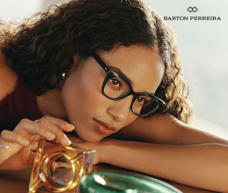 Adult female wearing green-framed eyeglasses rests her head, glancing towards the foreground, with decorative glass cubes in view.