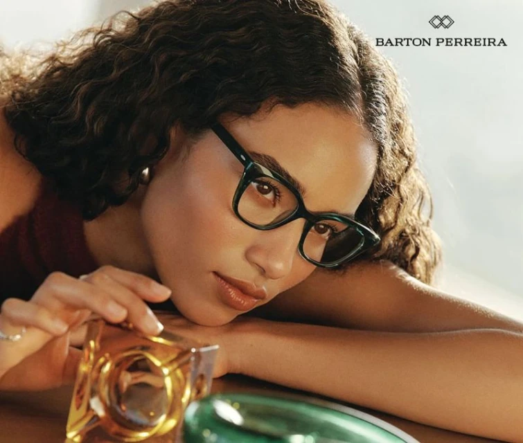 Adult female wearing green-framed eyeglasses rests her head, glancing towards the foreground, with decorative glass cubes in view.