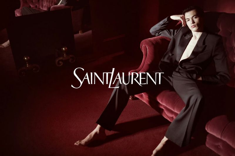 Man in black suit poses on red velvet chaise lounge in an indoor studio, showcasing Saint Laurent logo in a fashion-editorial setting.