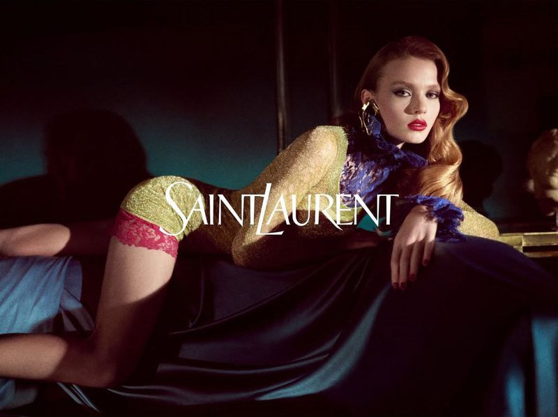 Fashion editorial shot of a young adult woman with red lipstick, laying on a dark blue satin chaise lounge wearing sheer gold top.