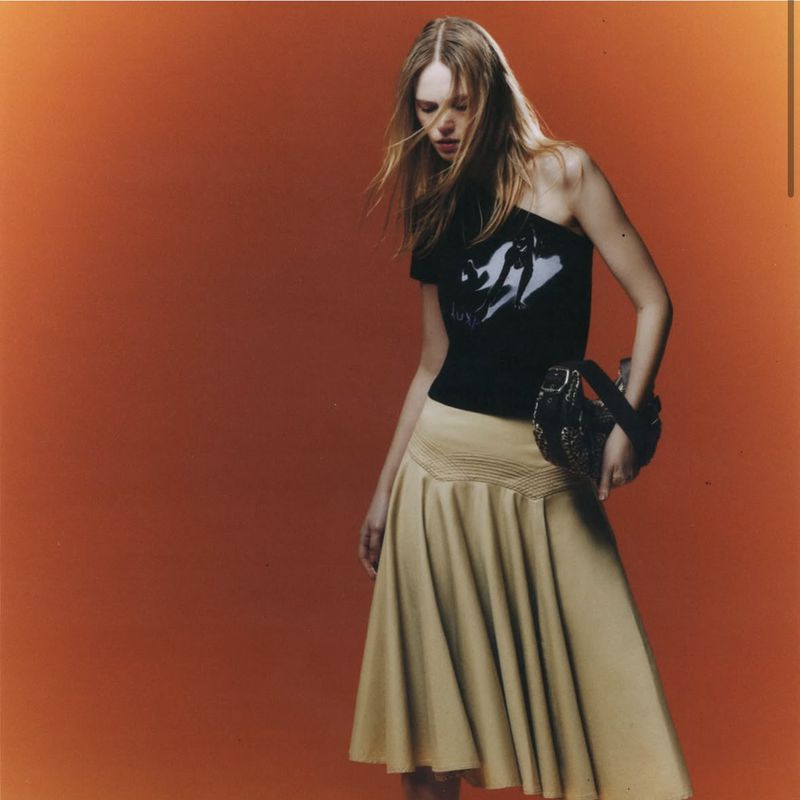 Fashion shot of a blonde woman in a black graphic top and tan skirt holding a studded handbag, posing against an orange background.
