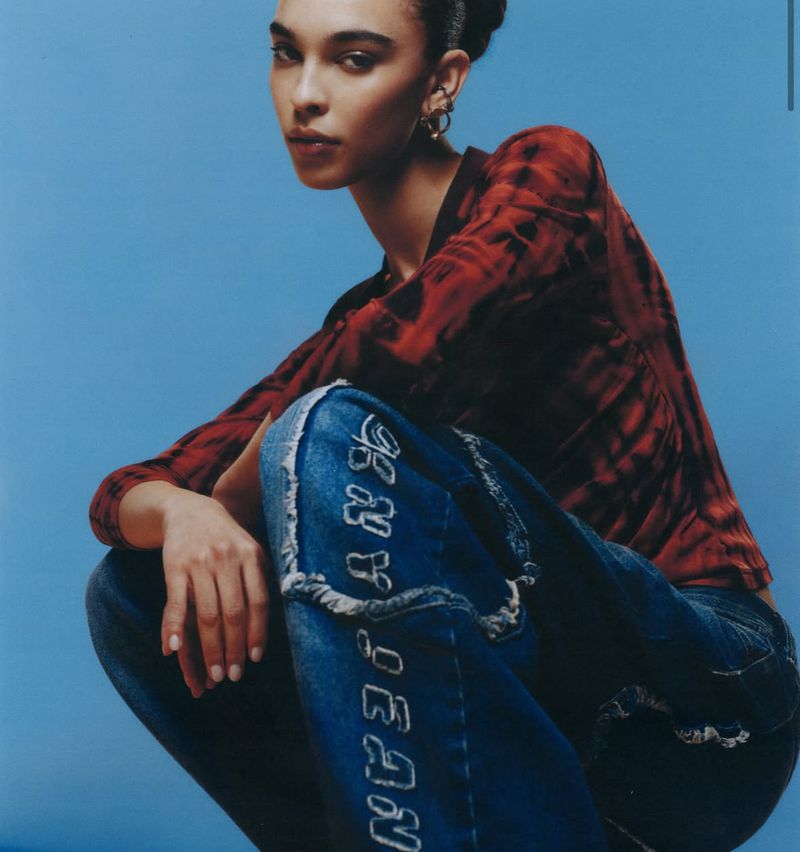 A woman poses in a studio environment, wearing a red and black tie-dye top with DKNY jeans, set against a blue backdrop for a fashion shot.