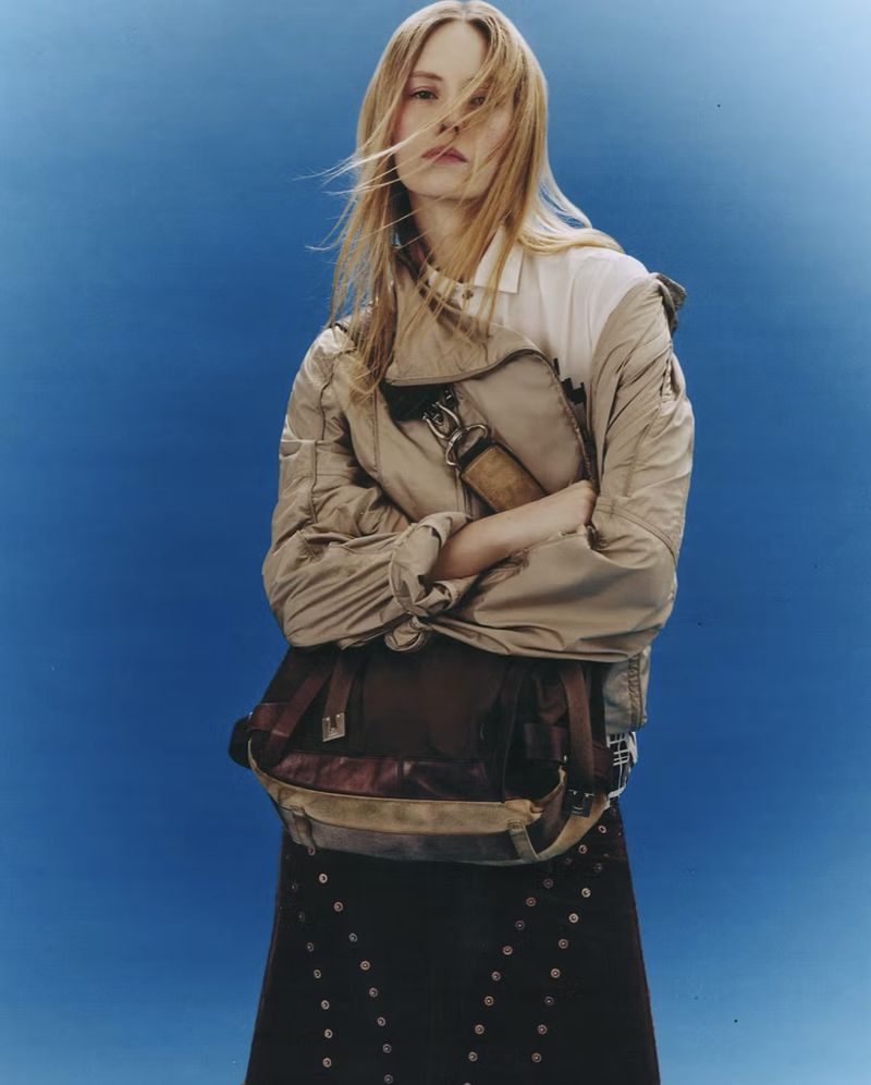 Model with long blonde hair wearing a beige jacket and holding a dark messenger bag against a blue backdrop.