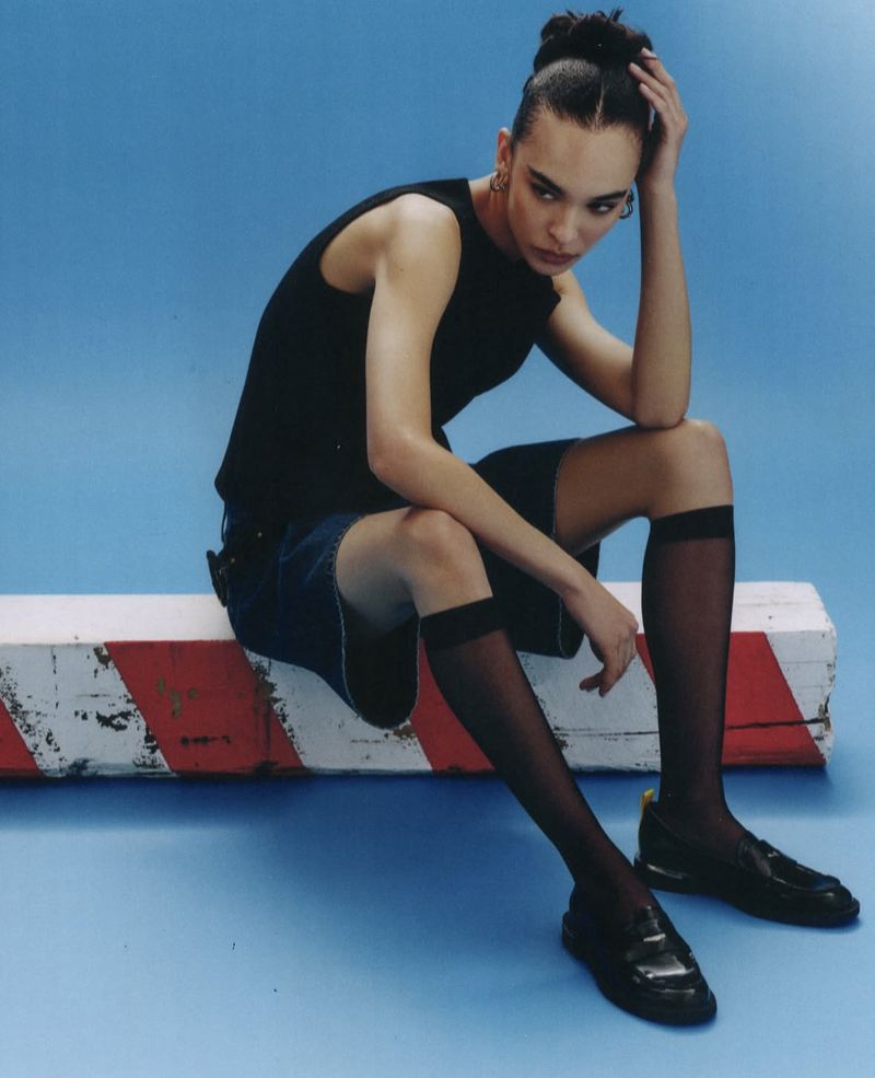 A woman with braided hair poses while sitting on a barrier, wearing a black top, denim shorts, knee-high socks, and platform loafers against a blue background.