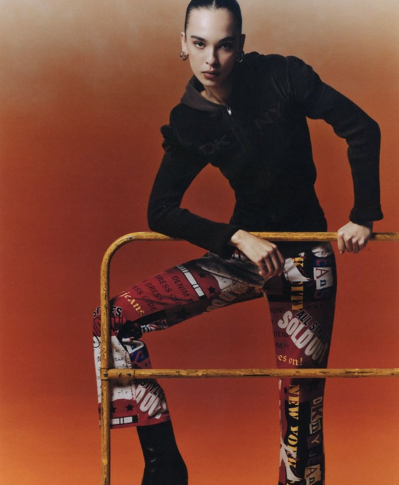 Fashion shot of a young adult female model posing with a yellow metal railing against an orange gradient backdrop, wearing patterned pants.