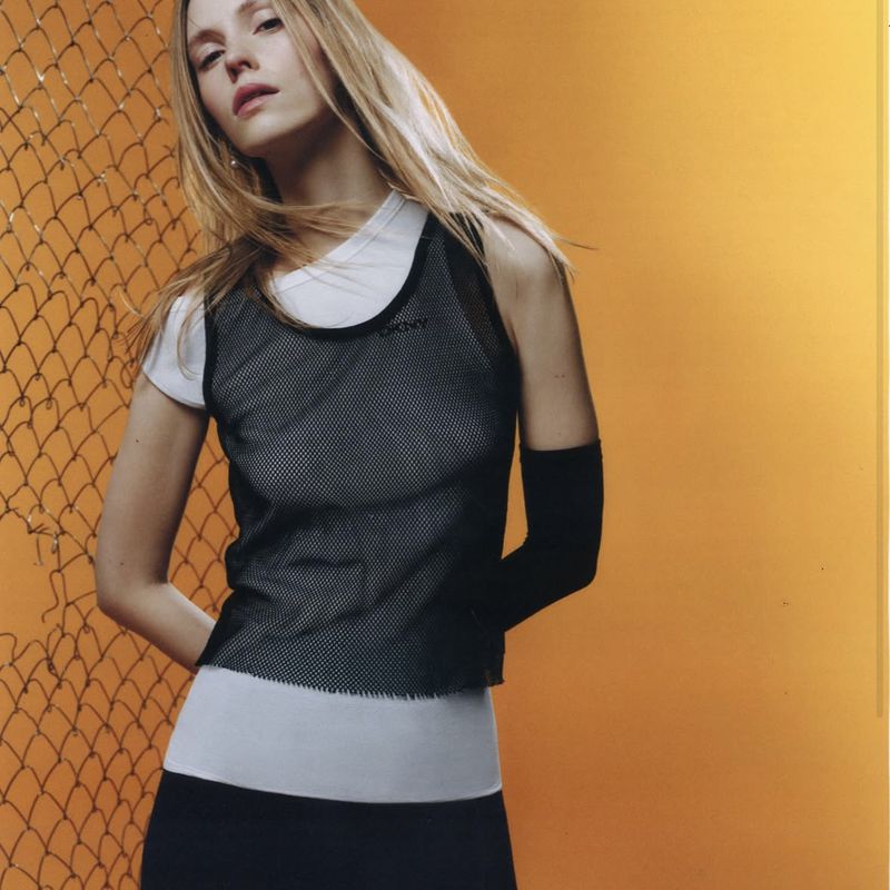 Model wearing a black mesh top over white layers against chain-link fence and orange wall
