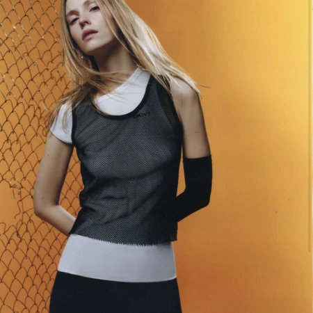 Model wearing a black mesh top over white layers against chain-link fence and orange wall
