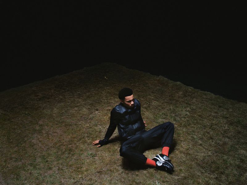 Man sitting on a grassy hilltop at night, wearing black puffer jacket, black pants and orange socks. The sky is dark.