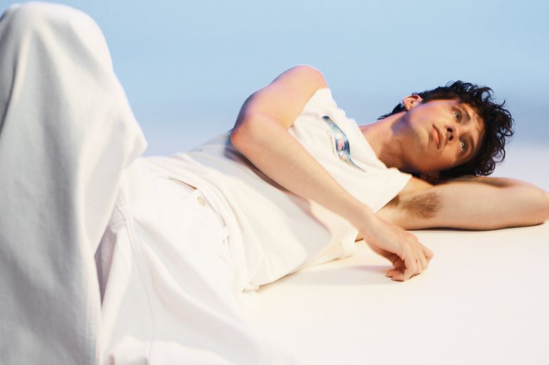 Young adult male lying on his side wearing white pants and a T-shirt in a photo studio with a light blue background. 
