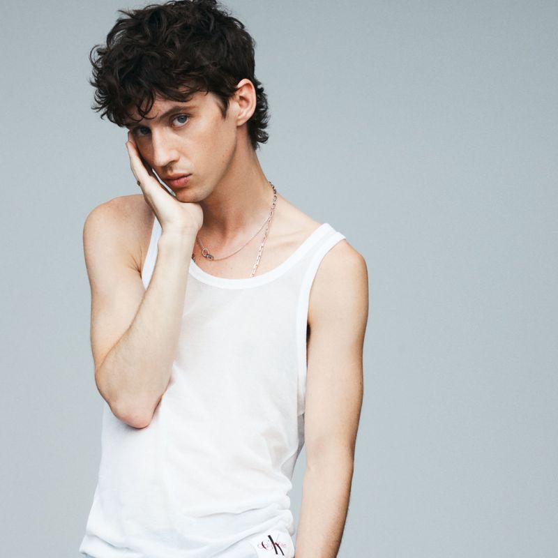 Troye Sivan wearing a white tank top with hand on cheek against a grey studio background for Russh I Am Love editorial
