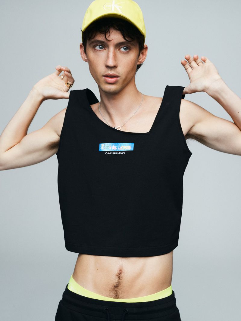 A young adult with light skin and dark hair wears a black crop top and yellow cap with the Calvin Klein Jeans logo, set against a gray background.