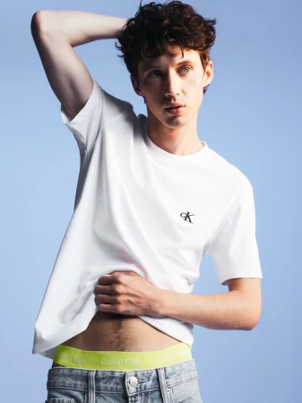 A young adult male poses in a studio environment, wearing a white t-shirt and jeans, with a light blue background, showcasing fashion.