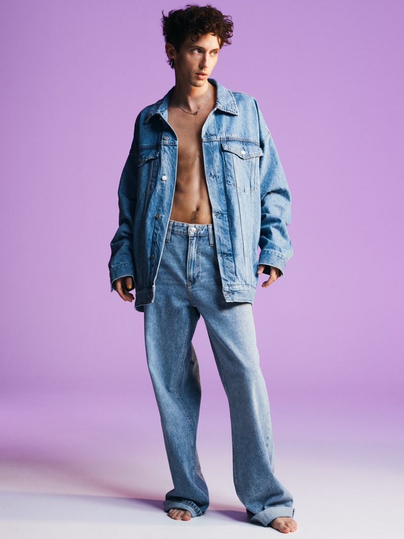 Full-length shot of a slender adult male, wearing a denim jacket and baggy jeans against a solid lavender background in a studio.