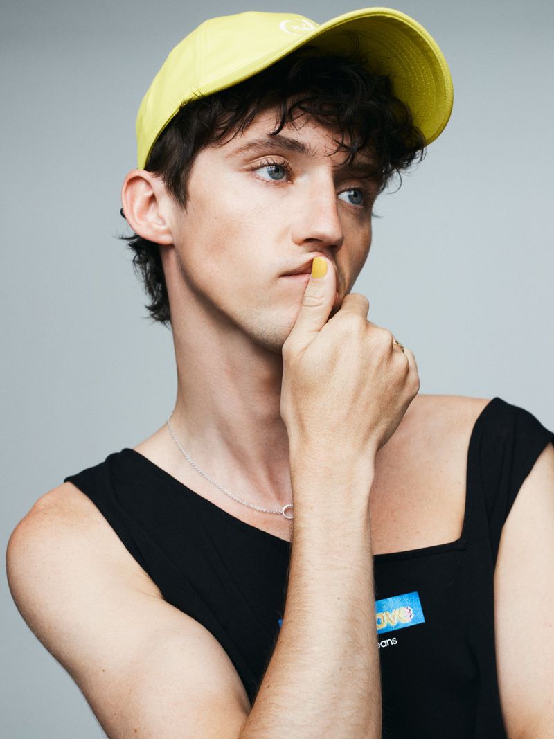 Portrait of a young adult male with dark hair wearing a yellow baseball cap and a black tank top in a photo studio setting.