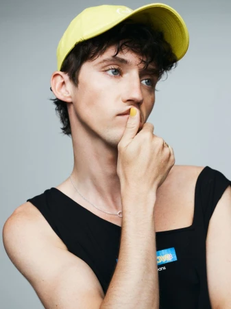 Portrait of a young adult male with dark hair wearing a yellow baseball cap and a black tank top in a photo studio setting.