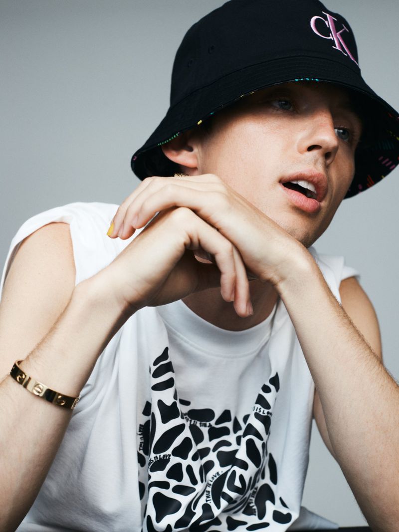 Troye Sivan wearing a black bucket hat and white graphic tank top against a plain grey backdrop for Russh magazine.