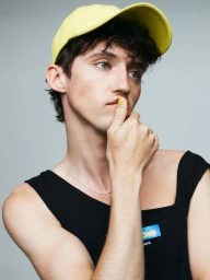 Troye Sivan wearing a yellow cap and black top with yellow nail polish against a grey studio backdrop