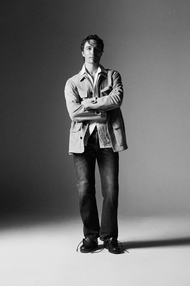Monochrome full-length portrait of a man standing with arms crossed against a plain grey studio backdrop.