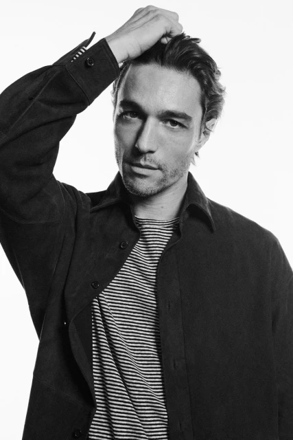 Black-and-white studio portrait of a young adult male with dark hair wearing an open collared dark shirt and striped T-shirt, hand on head.