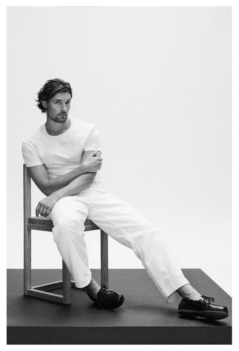 A man seated on a wooden chair wearing a white t-shirt, white trousers, and dark loafers against a plain light background.