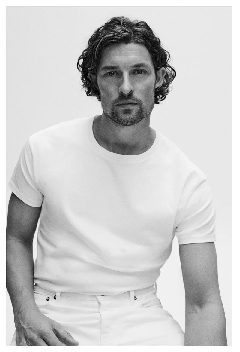 Monochrome portrait of a man with curly hair wearing a white t-shirt and jeans, posing in a photo studio with bright lighting.