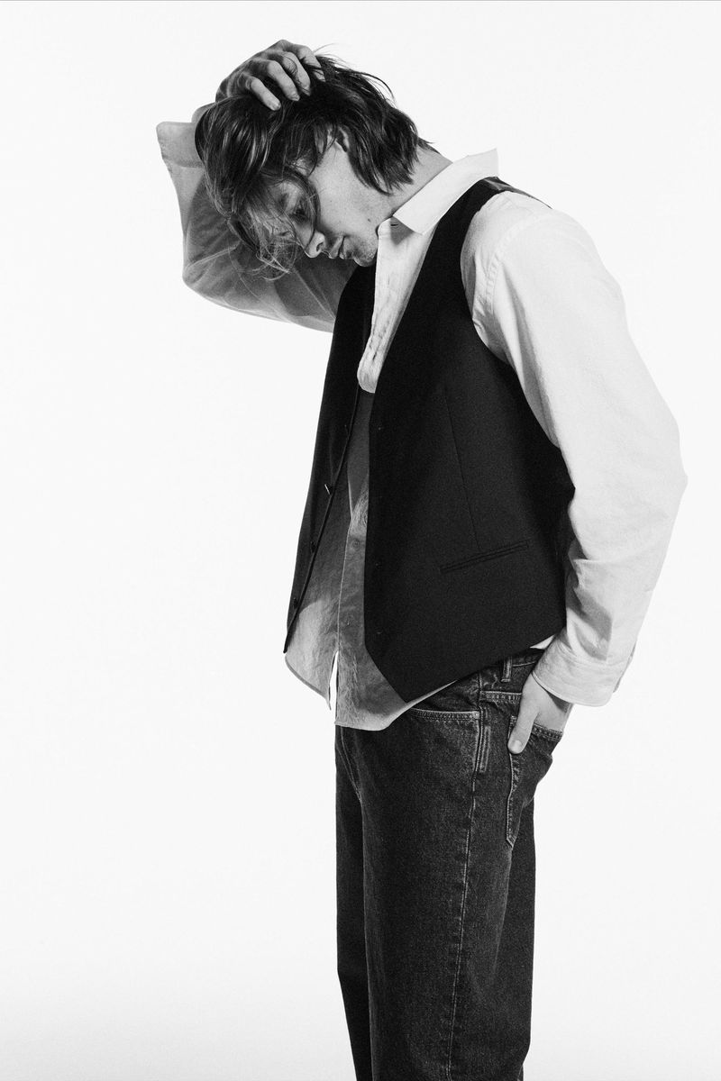 Black and white portrait of a young adult male in a vest, shirt, and jeans, with his hand on his head, against a white background.