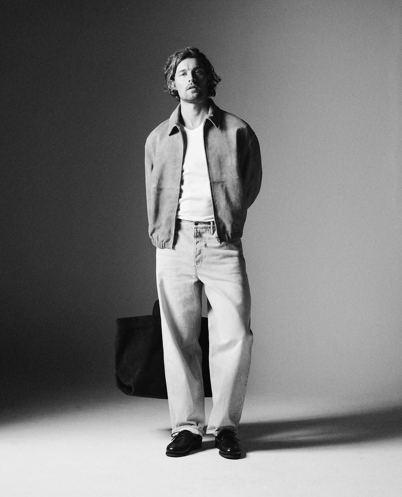 Full-length portrait of a male model wearing a casual jacket, white t-shirt, and light jeans against a grey studio backdrop.