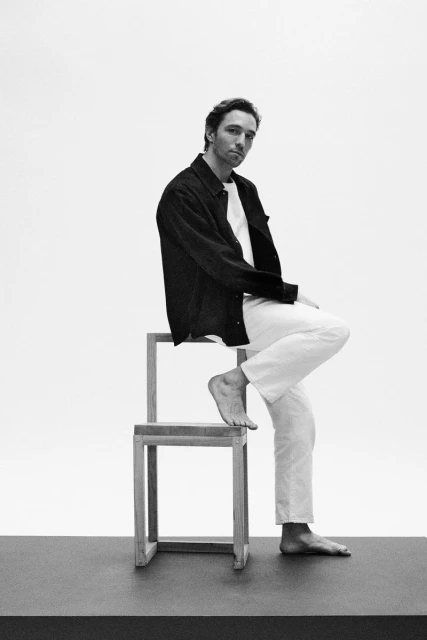 Black and white portrait of a man sitting barefoot on a wooden structure against a bright white backdrop in a photo studio.