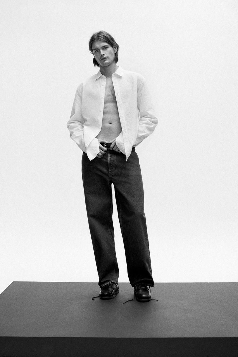 Monochrome studio shot of a slender male model in an open white linen shirt and dark jeans, standing on a black platform.