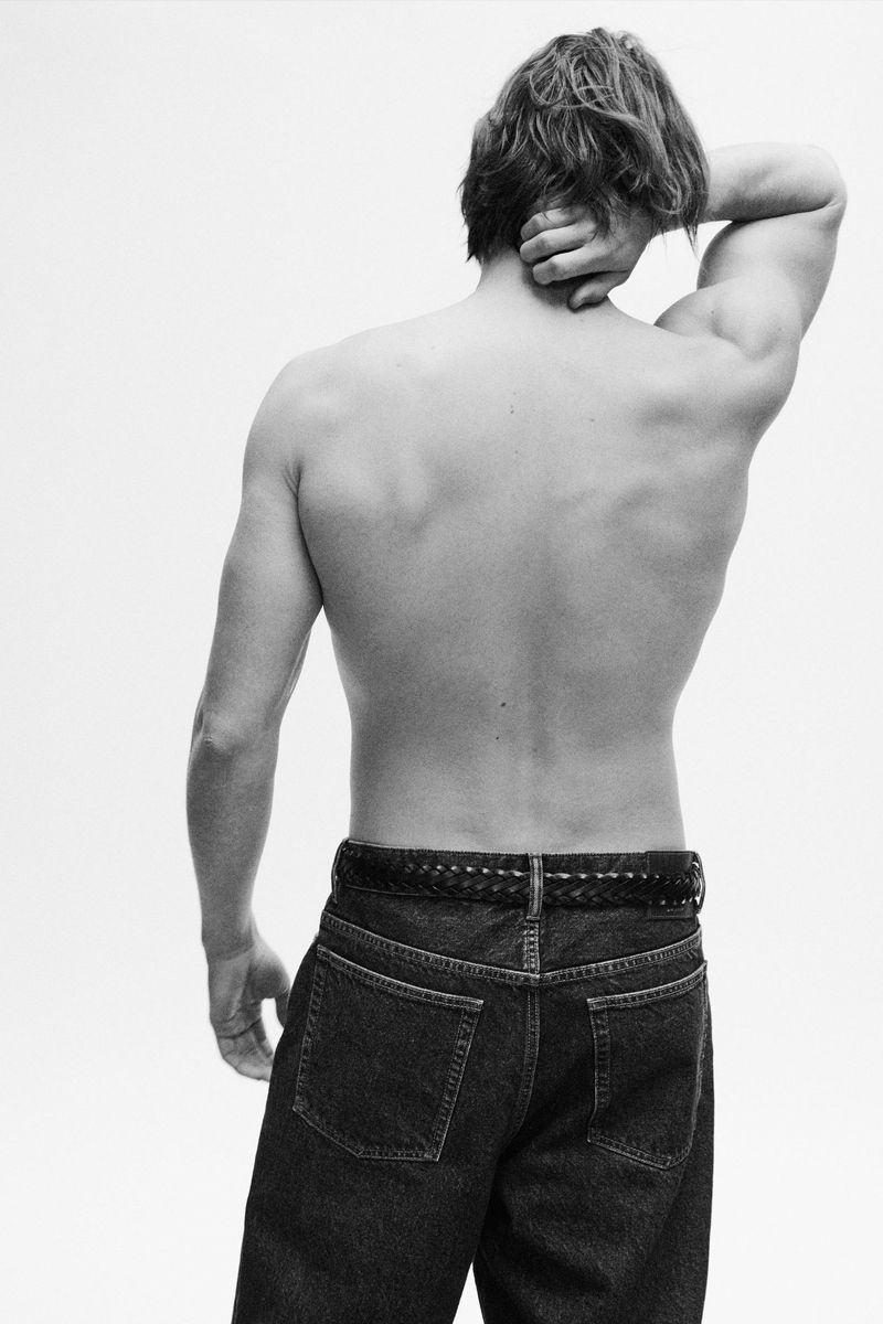 Black and white portrait of a shirtless adult male with blond hair wearing jeans and a belt, standing against a white background.