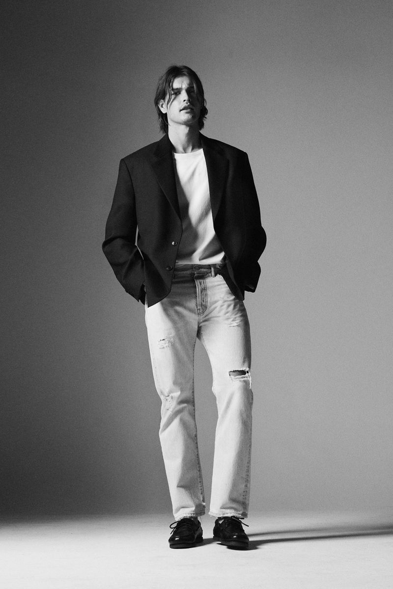 Man wearing a black blazer over a white t-shirt and light wash jeans standing against a grey studio backdrop.