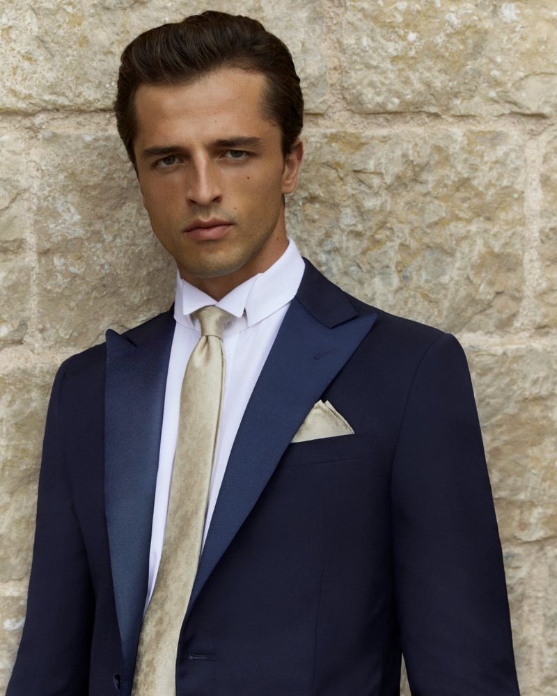 Elegant man in a navy blue tuxedo and champagne-colored tie poses against a stone wall in a fashion or formal wear portrait.
