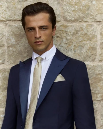 Elegant man in a navy blue tuxedo and champagne-colored tie poses against a stone wall in a fashion or formal wear portrait.