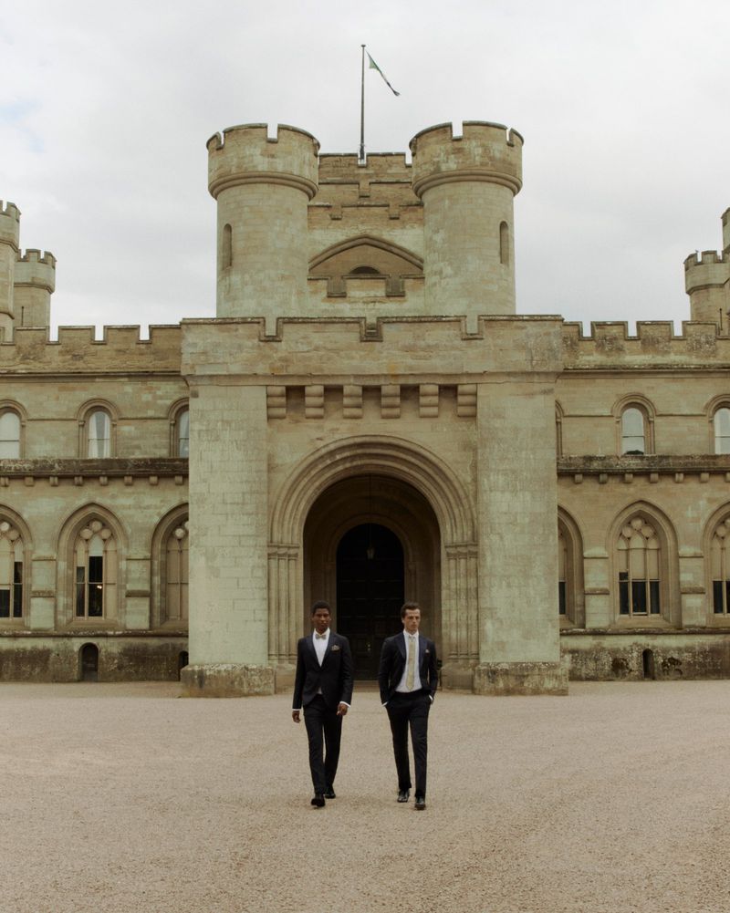 Two men in suits walk toward the camera in front of a large stone castle with towers, an arched entryway, and a flagpole.