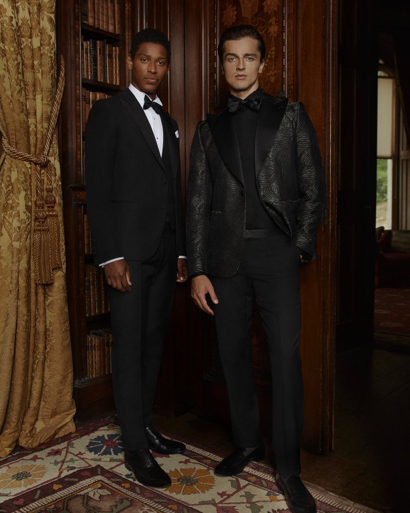 Two adult men in formal wear pose indoors. One wears a black tuxedo, and the other is in a patterned black jacket with a bow tie.