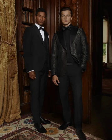 Two adult men in formal wear pose indoors. One wears a black tuxedo, and the other is in a patterned black jacket with a bow tie.