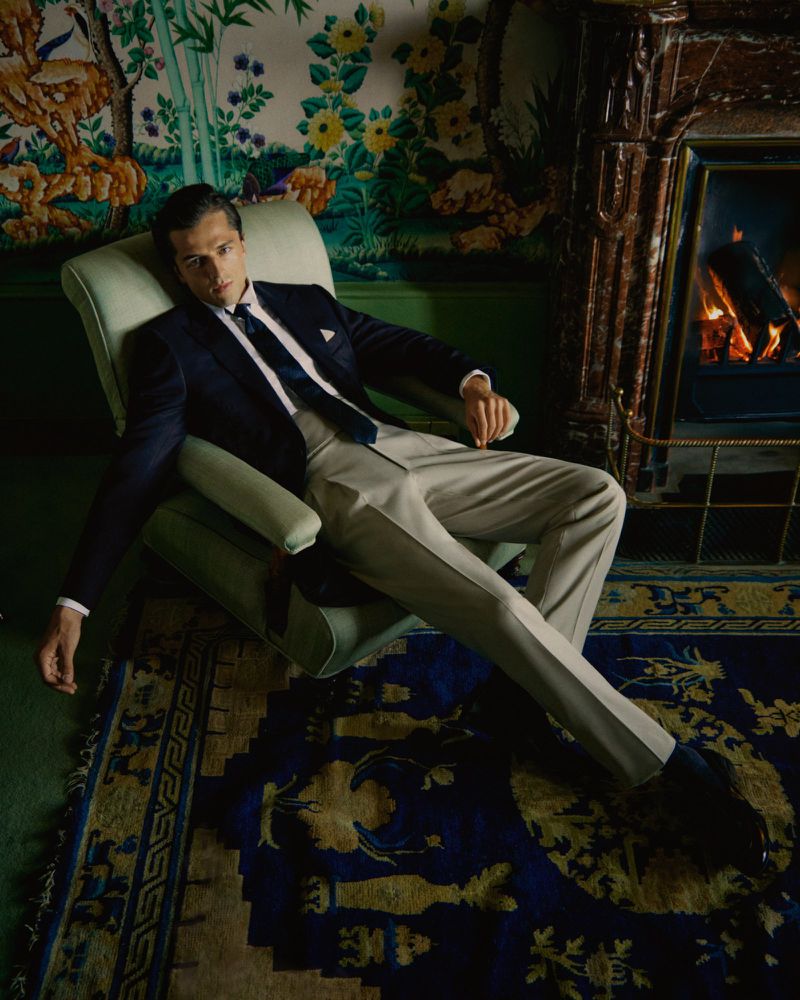 A man in a dark suit lounges in a light green armchair. He is wearing a tie and light-colored pants in an indoor sitting room.