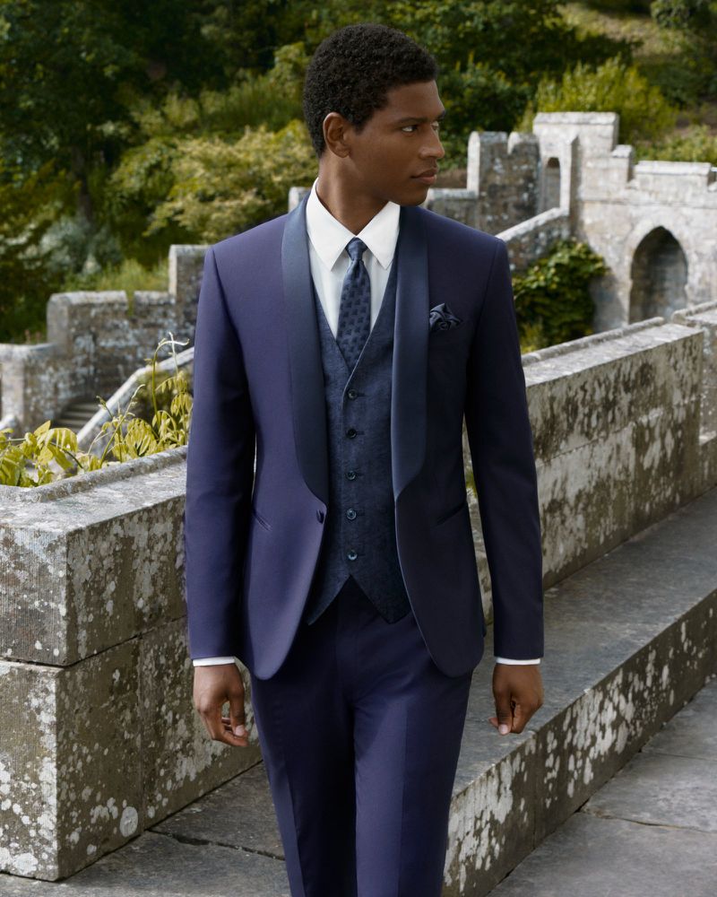 Dark-skinned male model in a blue suit with vest and tie walks along a stone wall with blurred green foliage in the background.